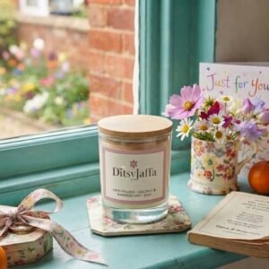 DitsyJaffa The Gift Collection scented candle jar sitting on a floral coaster on a teal windowsill, next to a matching floral mug filled with daisies, a gift box with a patterned ribbon, and a greeting card.
