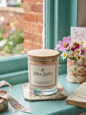 DitsyJaffa The Gift Collection scented candle jar sitting on a floral coaster on a teal windowsill, next to a matching floral mug filled with daisies, a gift box with a patterned ribbon, and a greeting card.